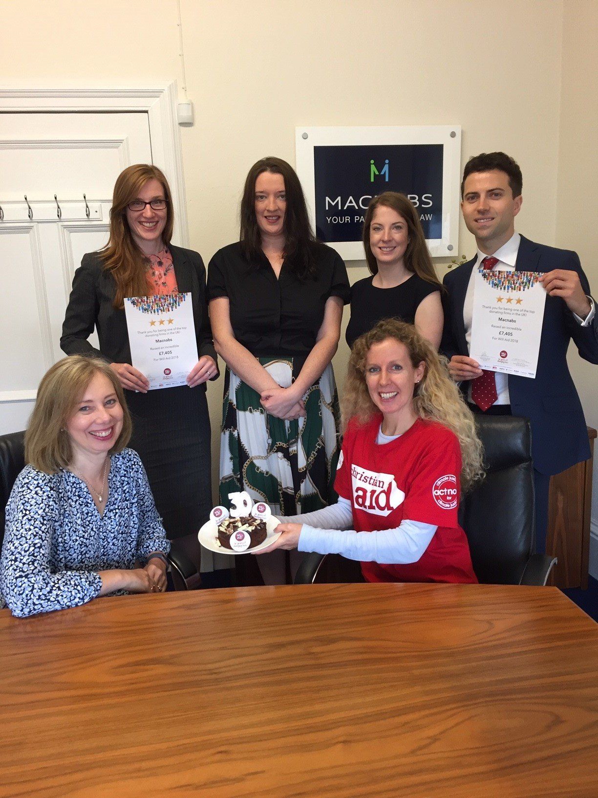 A group of people are sitting around a table holding certificates and a cake.