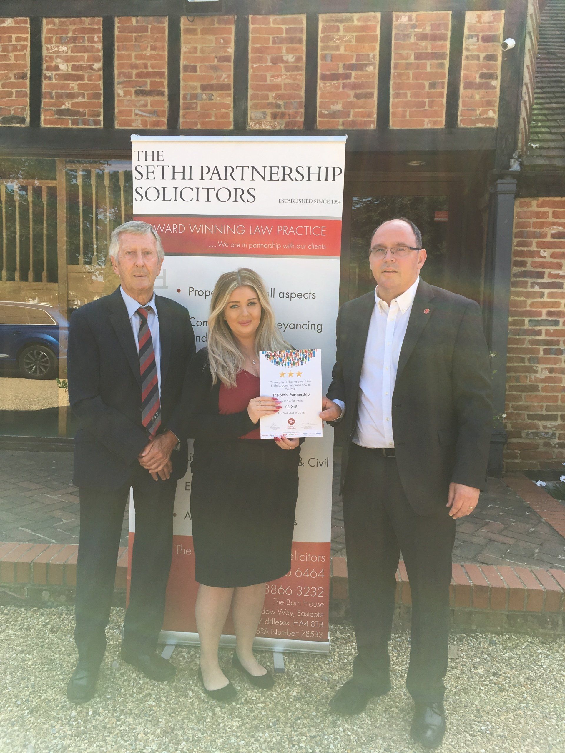 Three people standing in front of a brick building holding a certificate