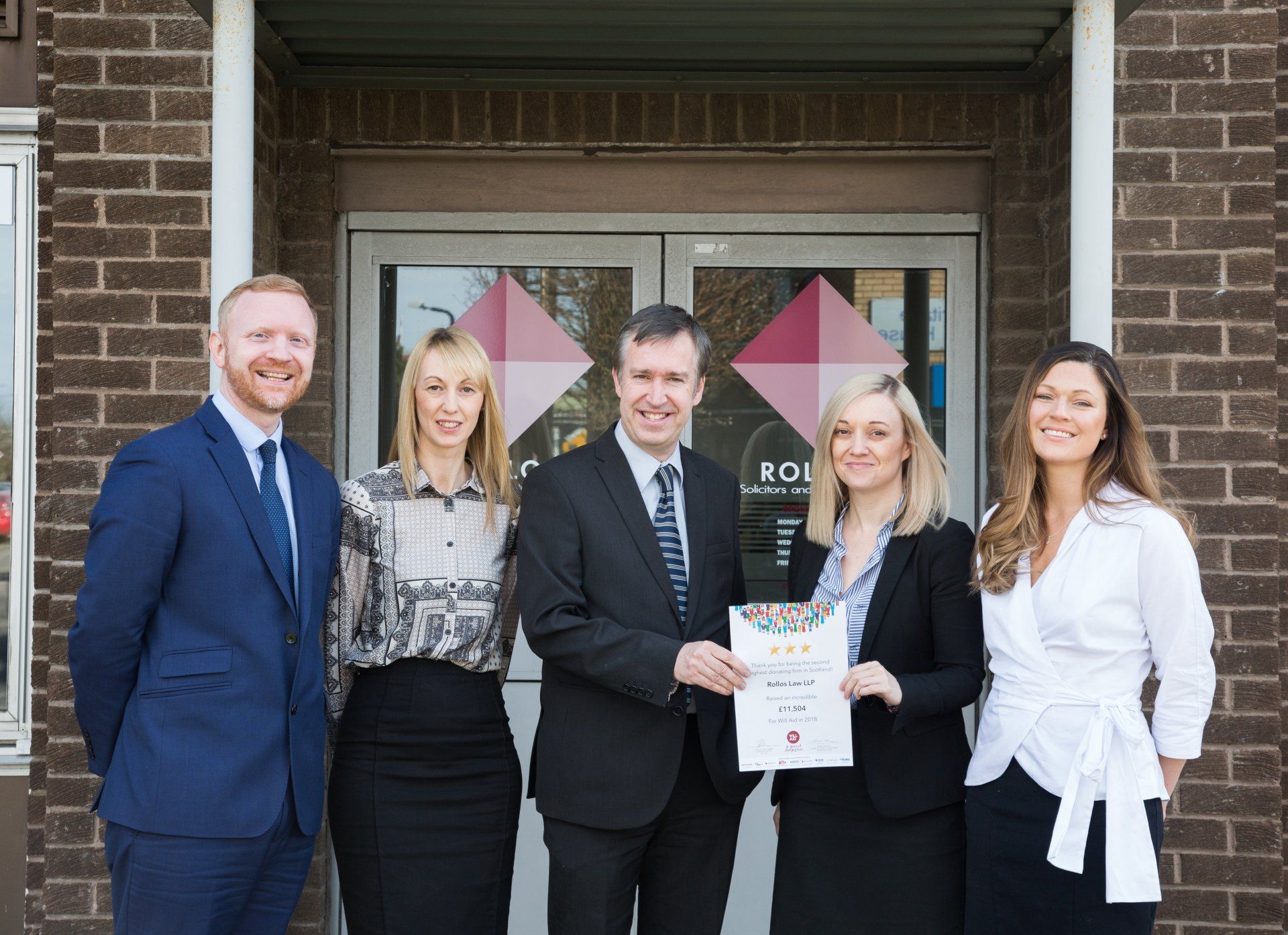 A group of people are standing in front of a brick building holding a certificate.