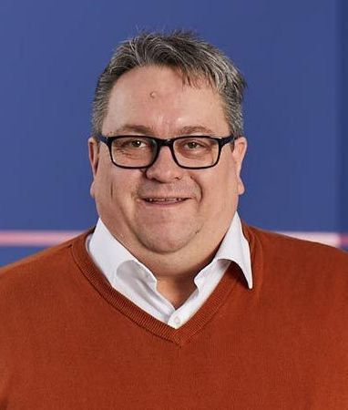 A man wearing glasses and an orange sweater is smiling for the camera.