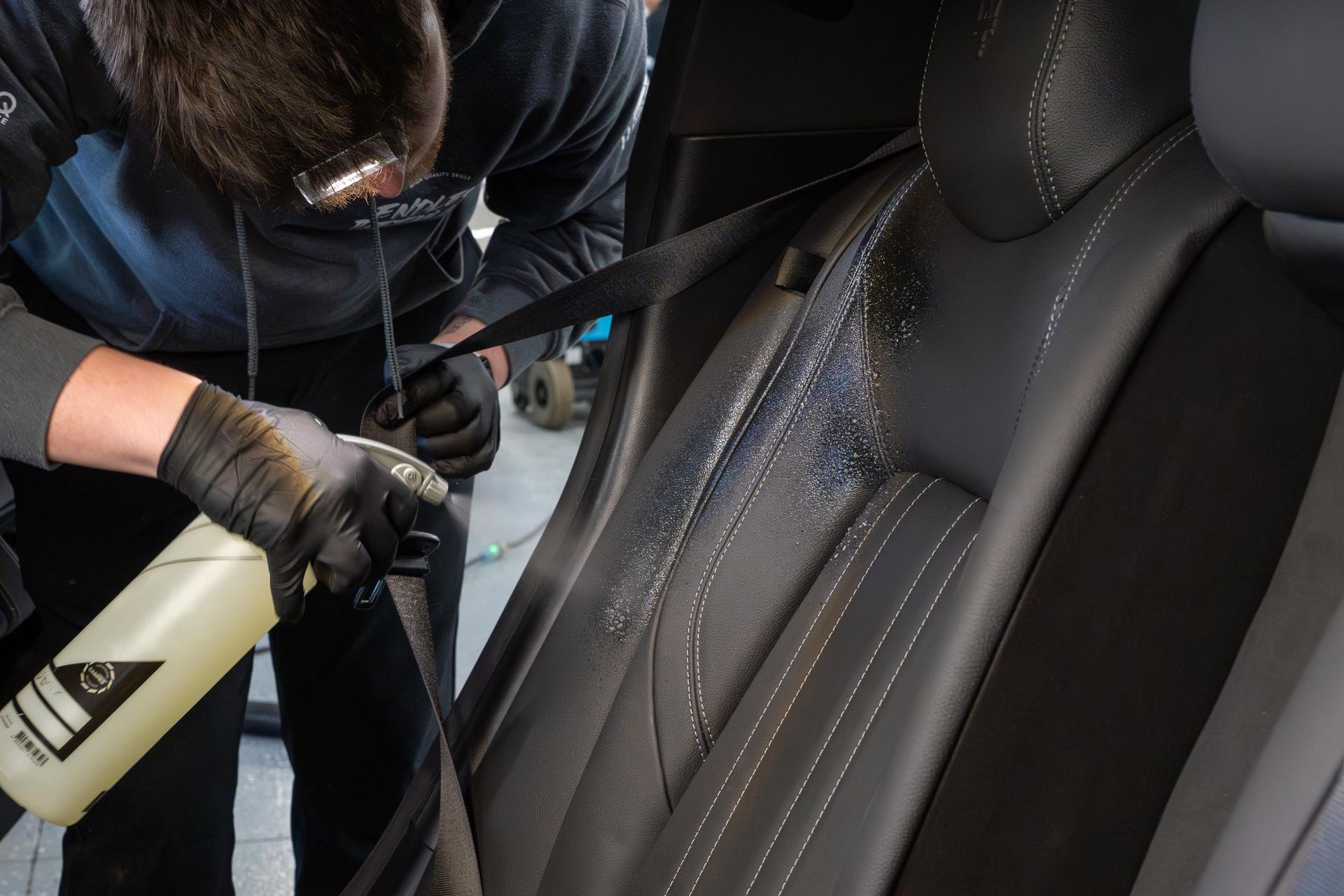 Person cleaning a black car seat with a spray bottle and wearing black gloves.