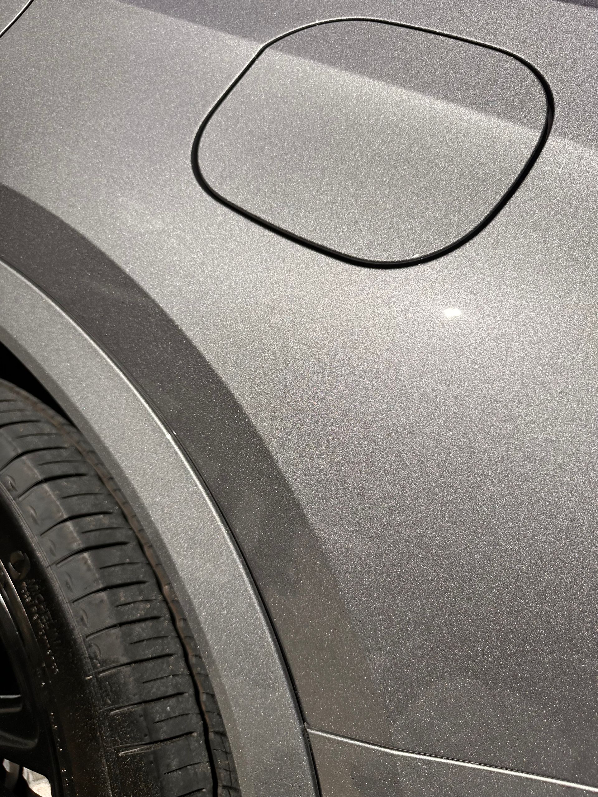 Close-up of a silver car's rear quarter panel, fuel door, and tire. Glitter-like texture visible on the car's surface.