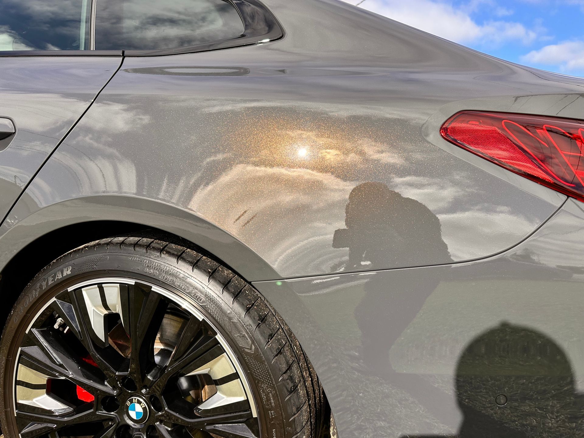 A gray BMW car's rear quarter panel with a reflective, bronze-colored shimmer, near a tire and a taillight, with a blue sky background.