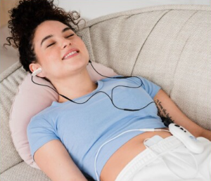A person with curly hair reclines on a couch with eyes closed, wearing earphones and using a white handheld device.