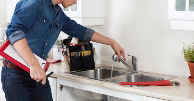 A man is fixing a sink in a kitchen while holding a clipboard.