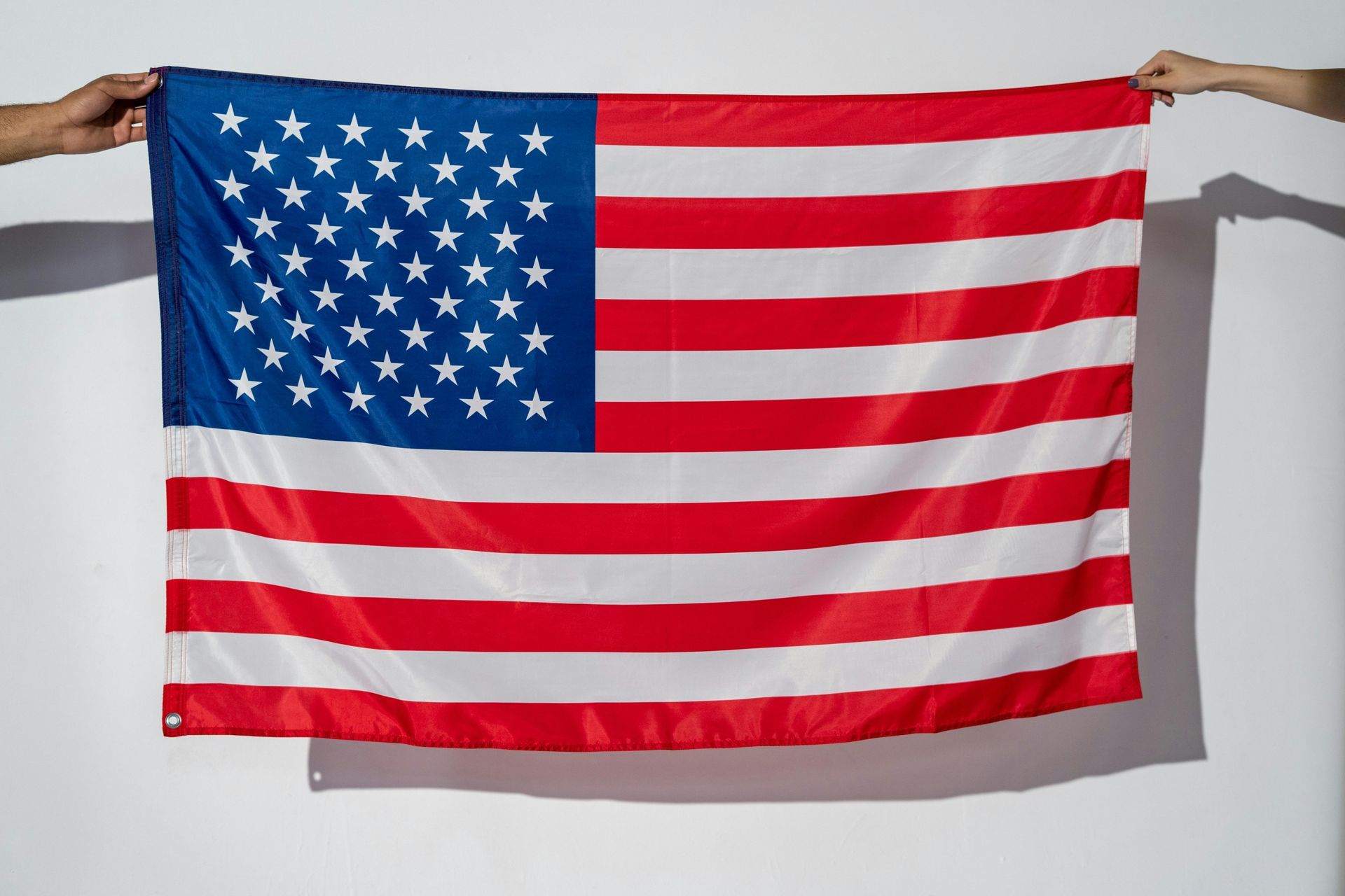 Two people are holding an american flag against a white wall.