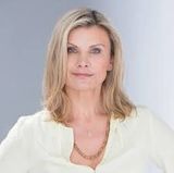 Eine Frau mit blondem Haar trägt ein weißes Hemd und eine goldene Halskette.