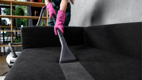 A woman is cleaning a black couch with a vacuum cleaner.