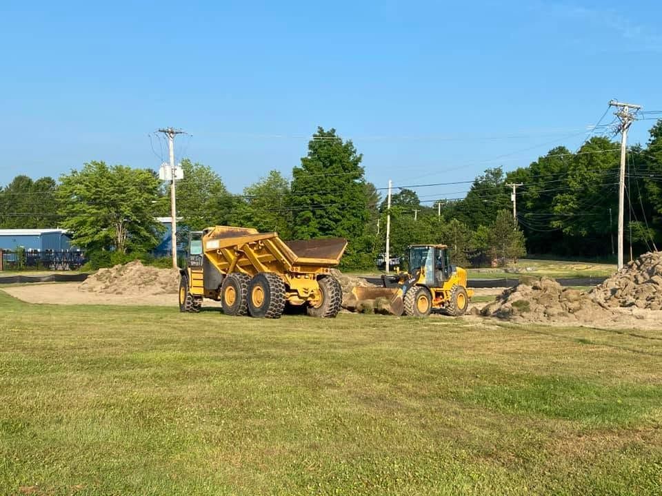 Yellow construction truck and backhoe working on a dirt pile in a grassy field under a blue sky