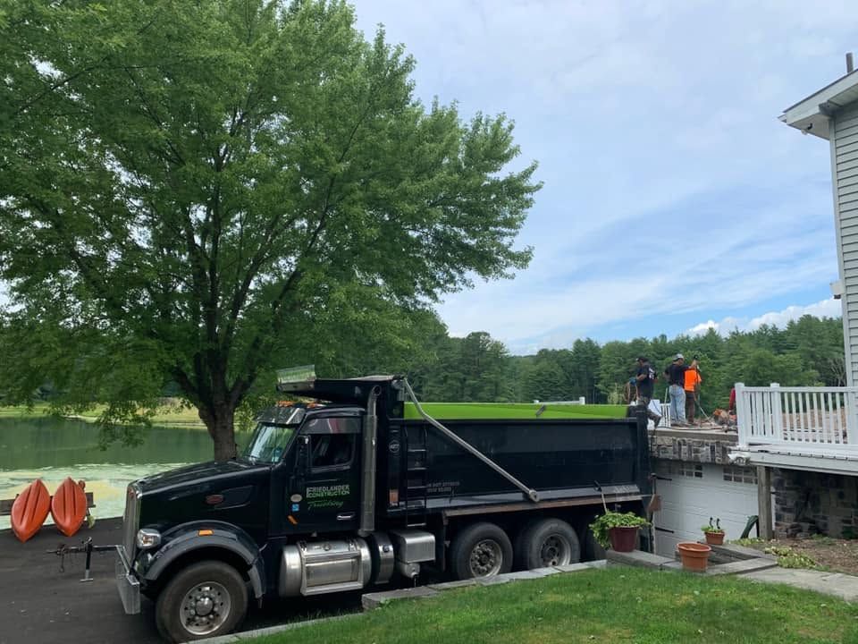 Black dump truck parked beside a house, with workers on the deck and a large tree nearby.