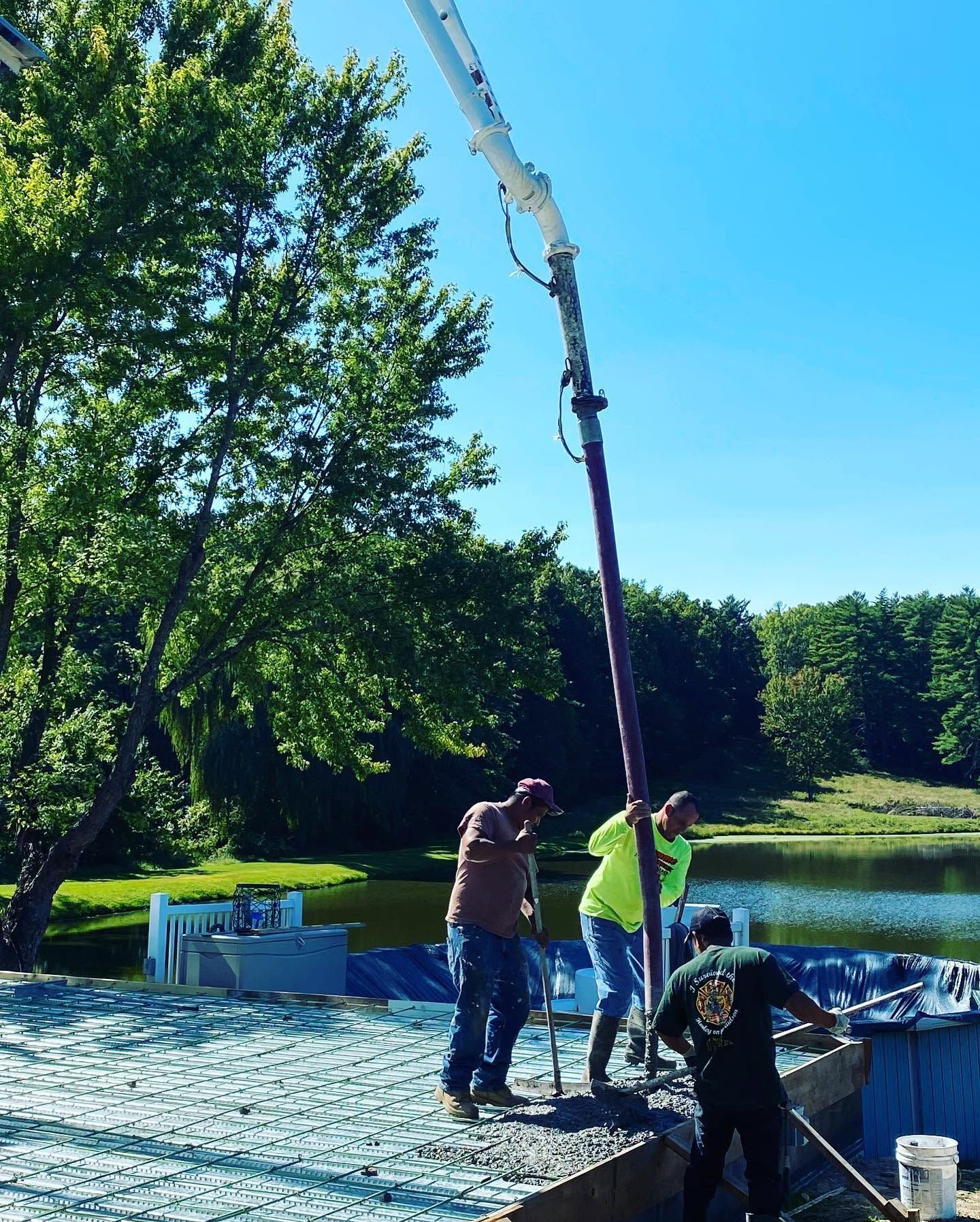 Workers operating a crane over a pond near a wooded shoreline on a sunny day