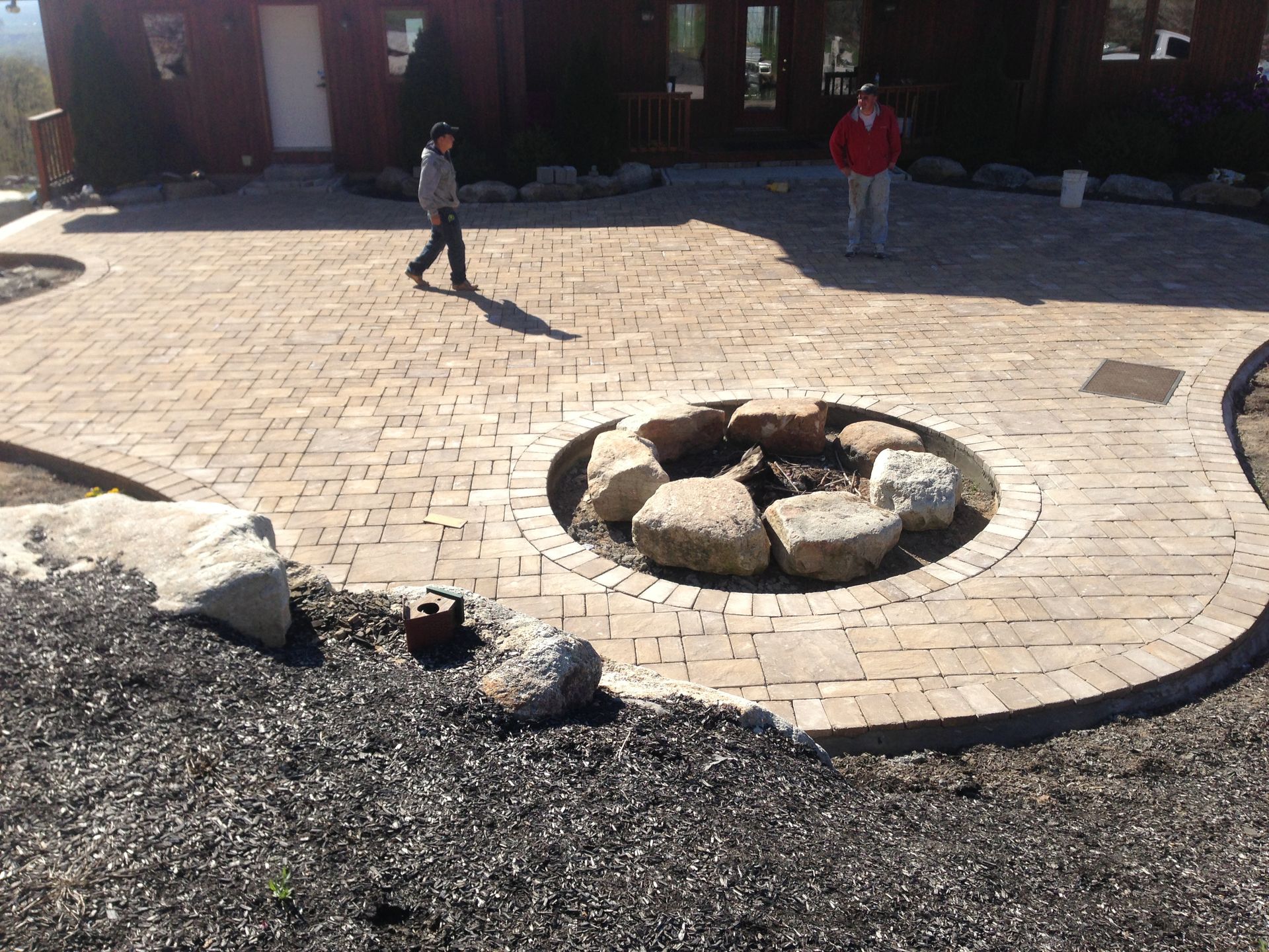 Stone fire pit in a sunlit courtyard with two people walking nearby and a building in the background