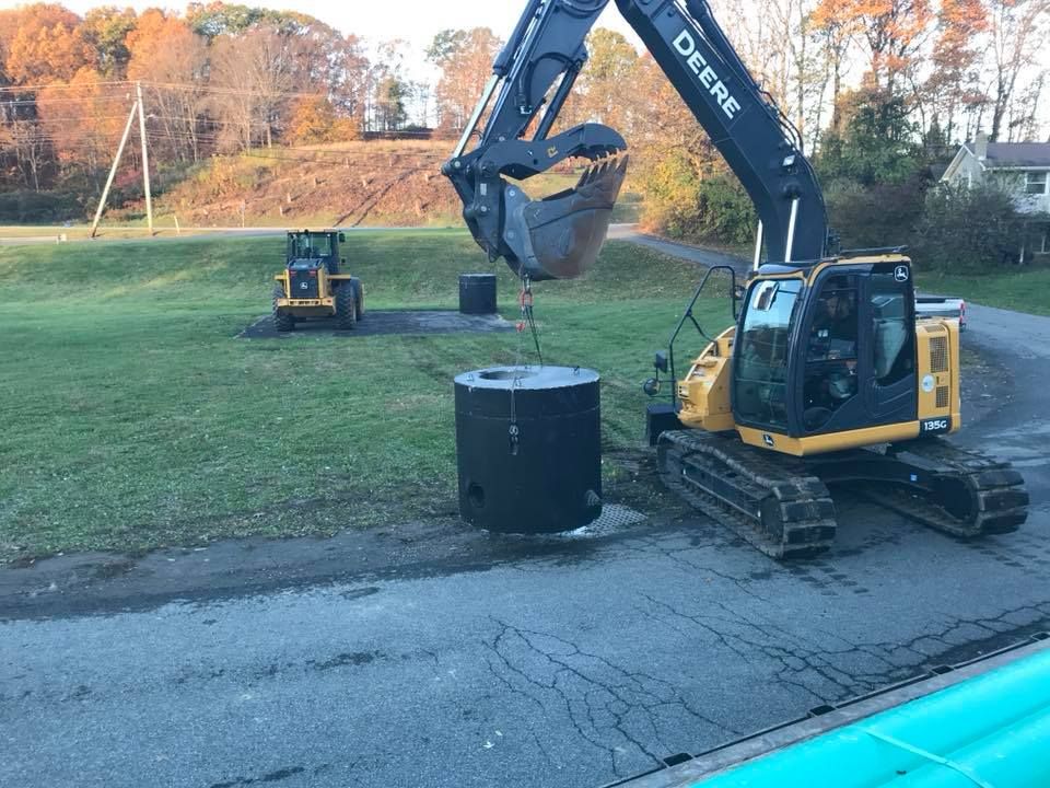 John Deere excavator lifting a black cylindrical tank on a grassy roadside near a road crack