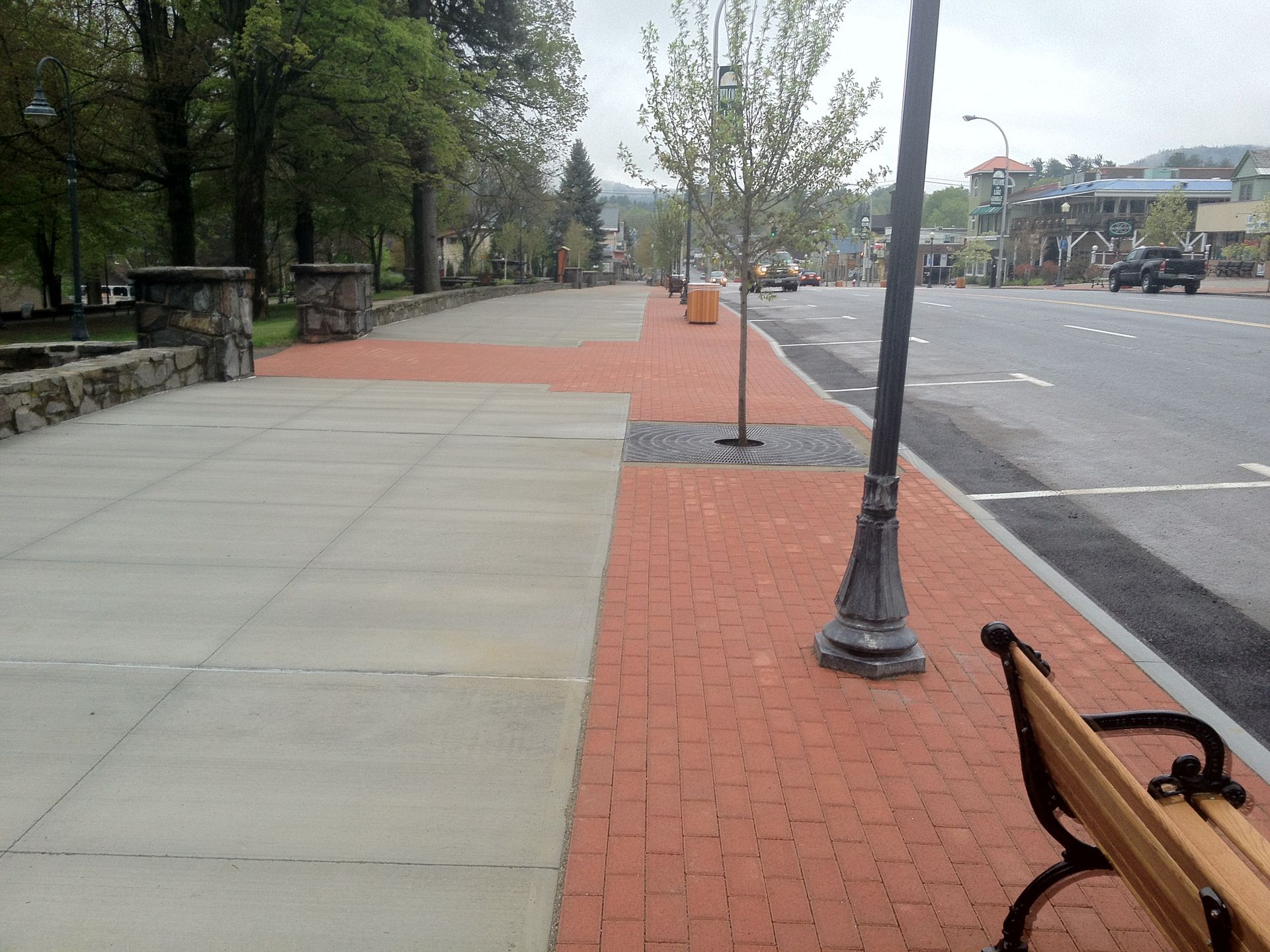 Empty sidewalk with red brick path, trees, lamp posts, and a road beside a park-like street.