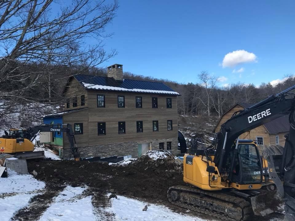 Construction excavator beside a two-story house in a snowy, wooded area