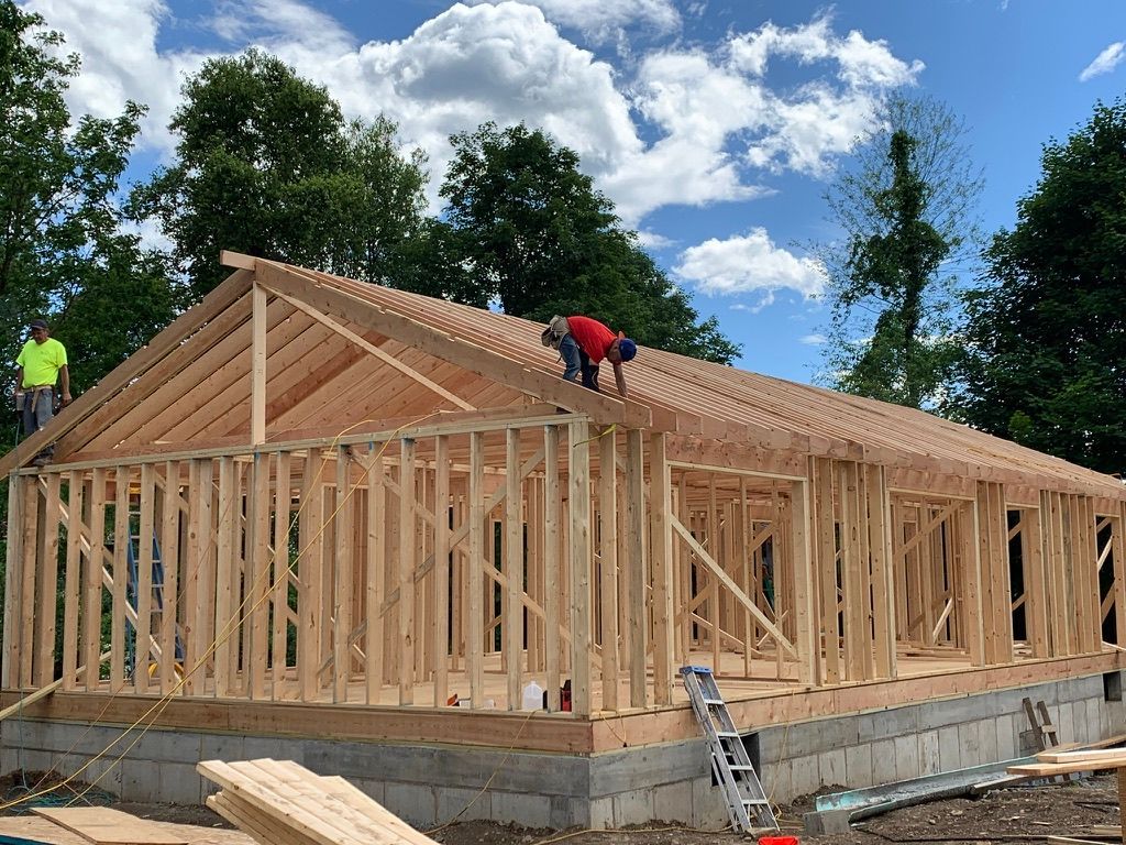 Wood-framed house under construction with workers on the roof and a ladder against the foundation.
