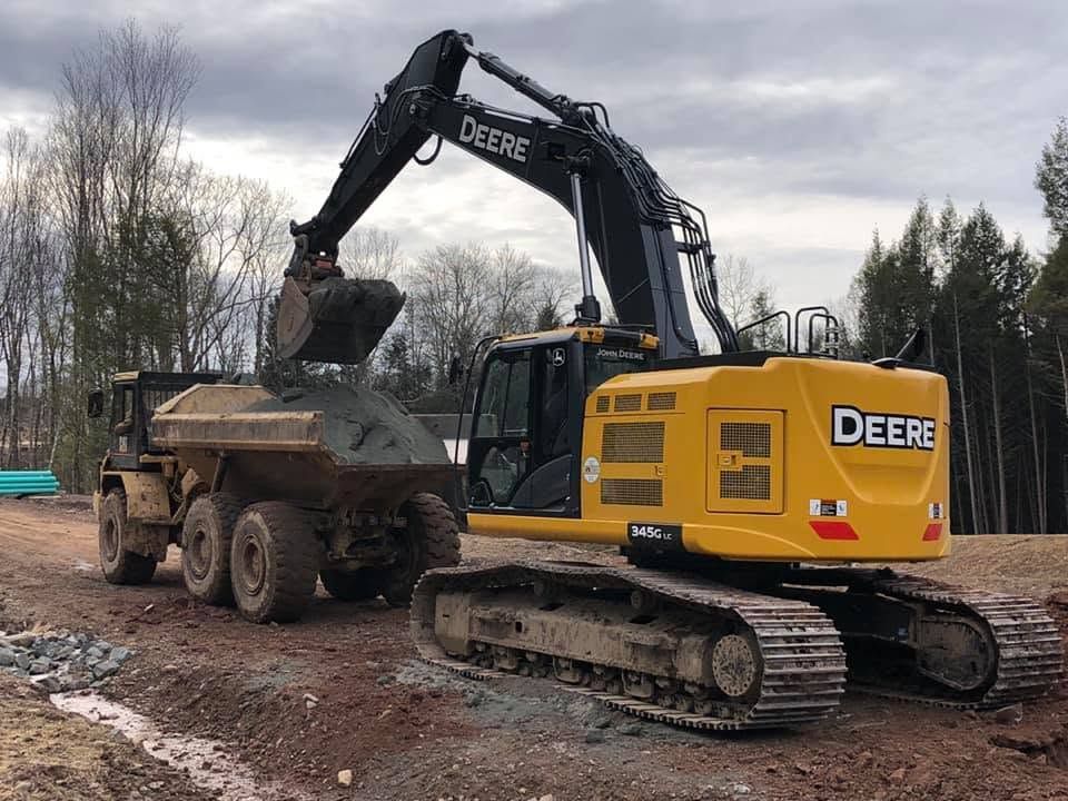 Yellow John Deere excavator loading dirt into a dump truck at a muddy construction site.