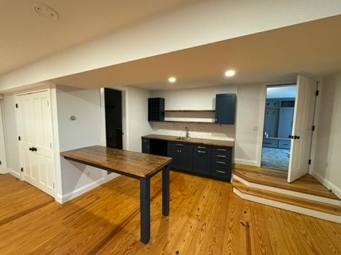 Empty kitchen with dark cabinets, wood floors, and a small wooden table in a bright room.