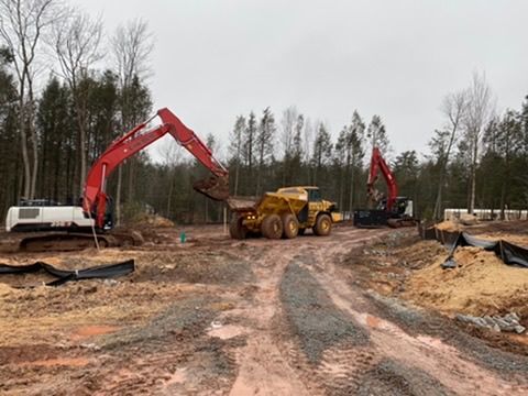 Excavators and a dump truck work on a muddy construction site near a wooded area.