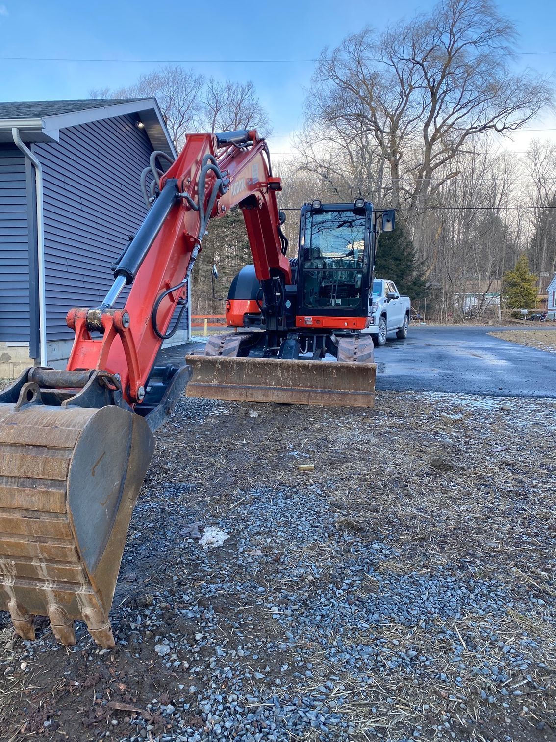 Red excavator parked beside a house on a gravel driveway with a blue truck in the background