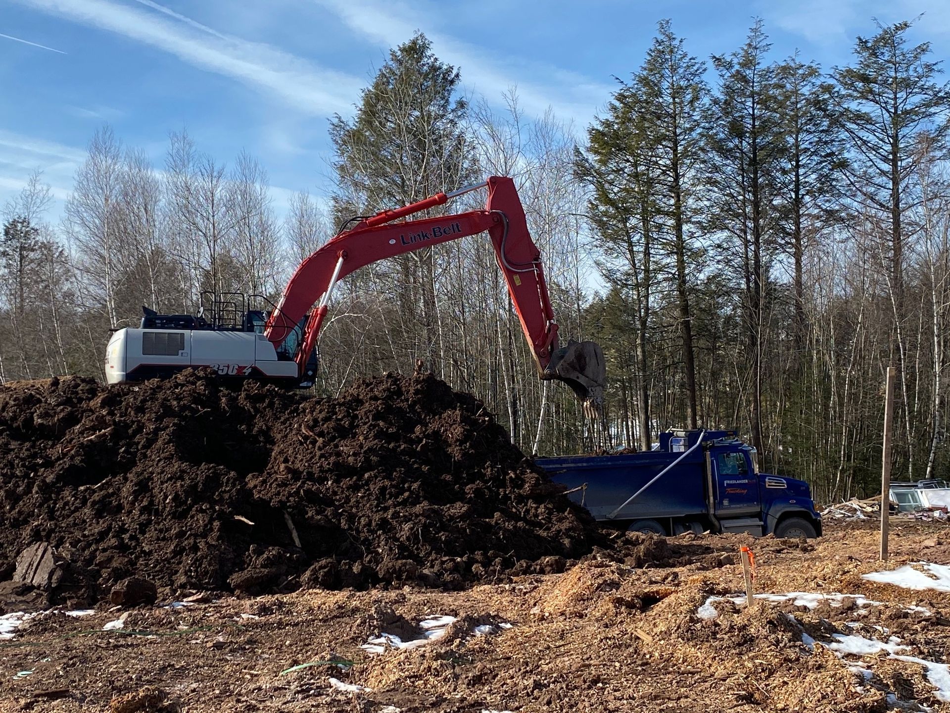 Red excavator loading dark soil into a blue dump truck in a snowy wooded clearing.