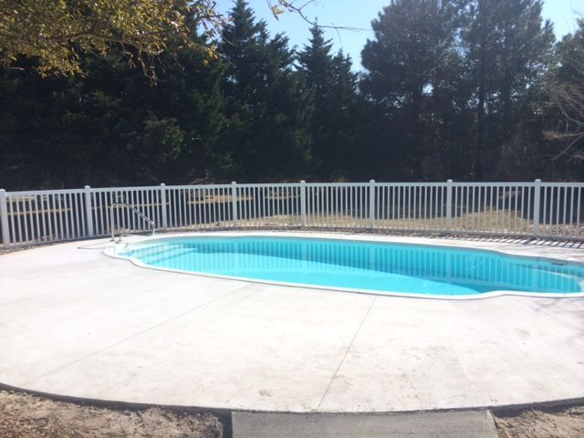 Outdoor swimming pool with white concrete deck and fence, surrounded by trees in daylight