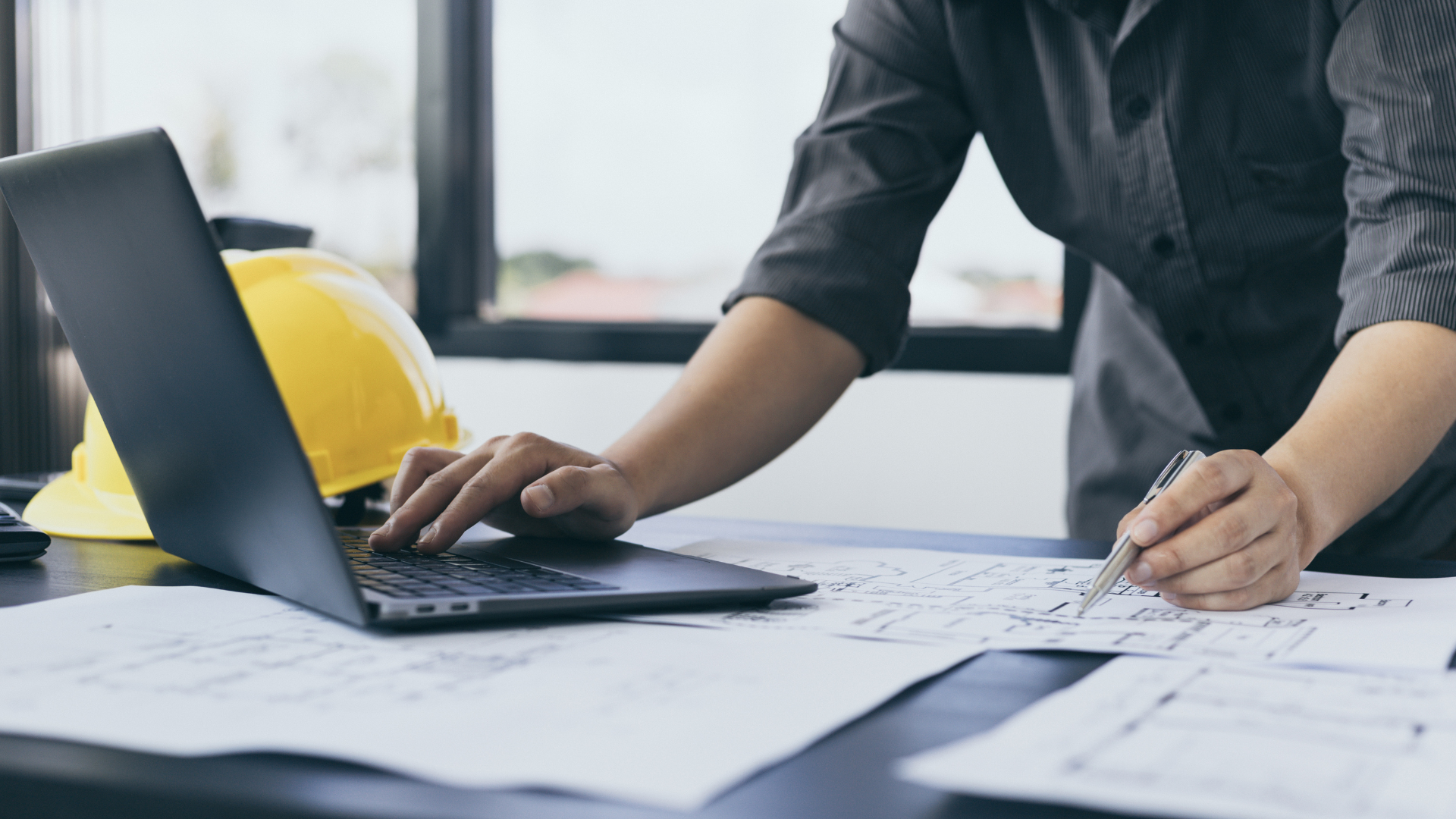 Person working on a laptop beside blueprints, with a yellow hard hat on the desk.