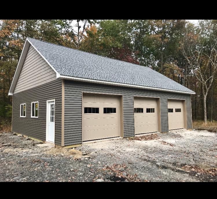 Gray three-bay garage with shingled roof on a gravel lot near trees.