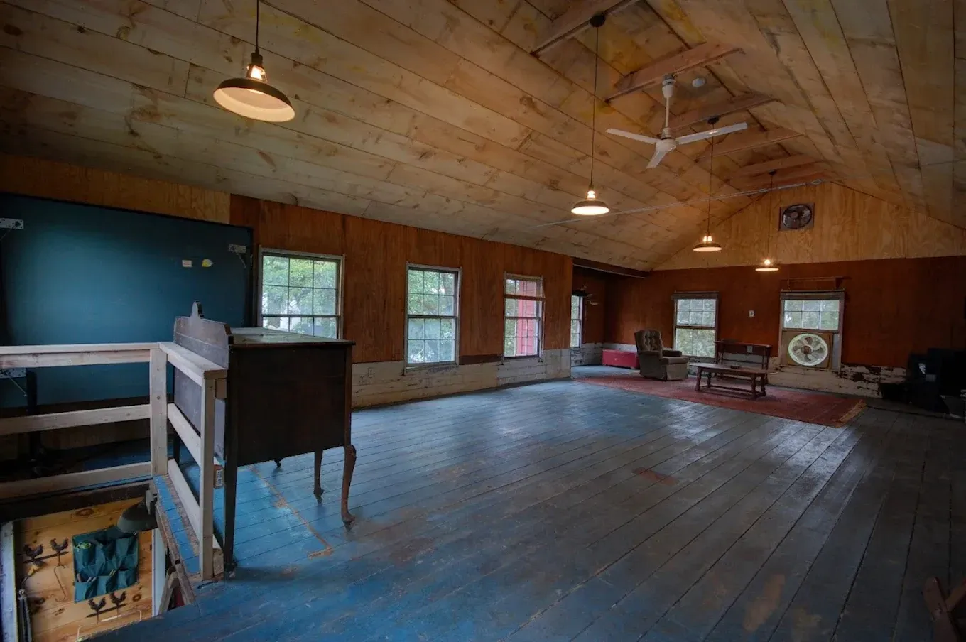 Empty rustic loft with wood-paneled walls, sloped ceiling, hanging lights, and windows overlooking dark wooden floors