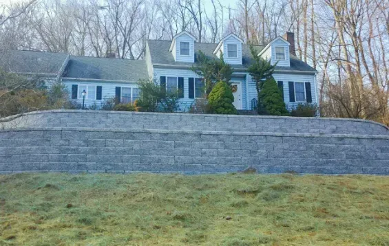 Blue house with dormer windows behind a stone retaining wall on a grassy hillside