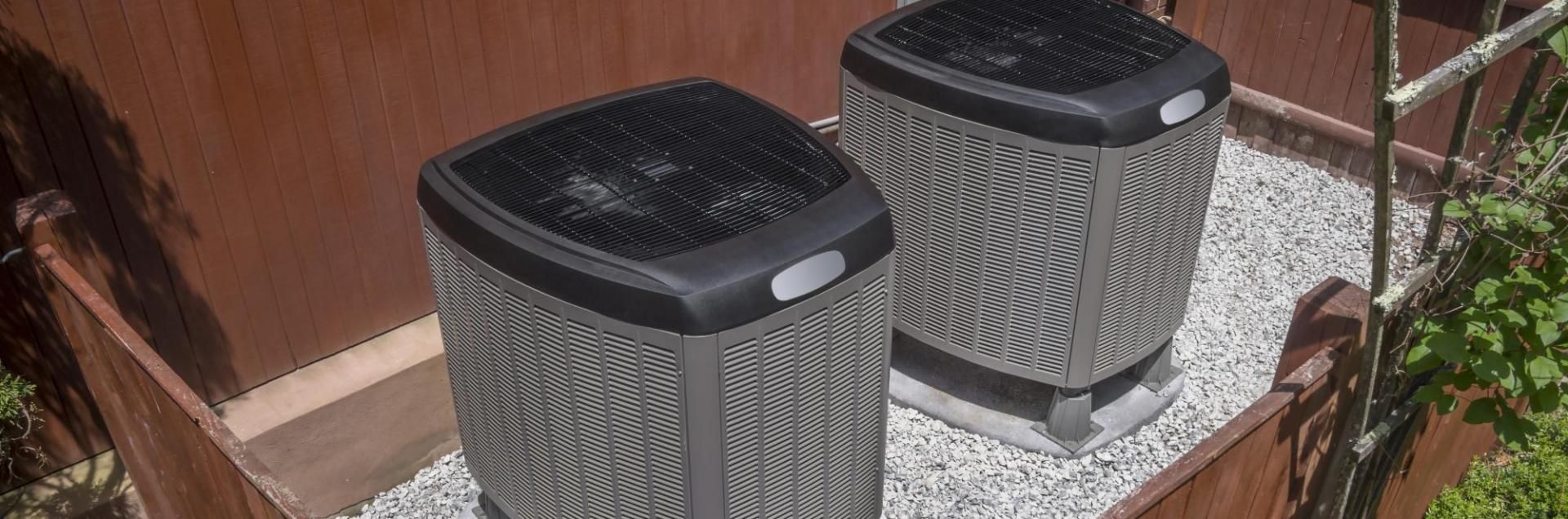 Two gray outdoor air conditioning units on gravel beside a wooden fence