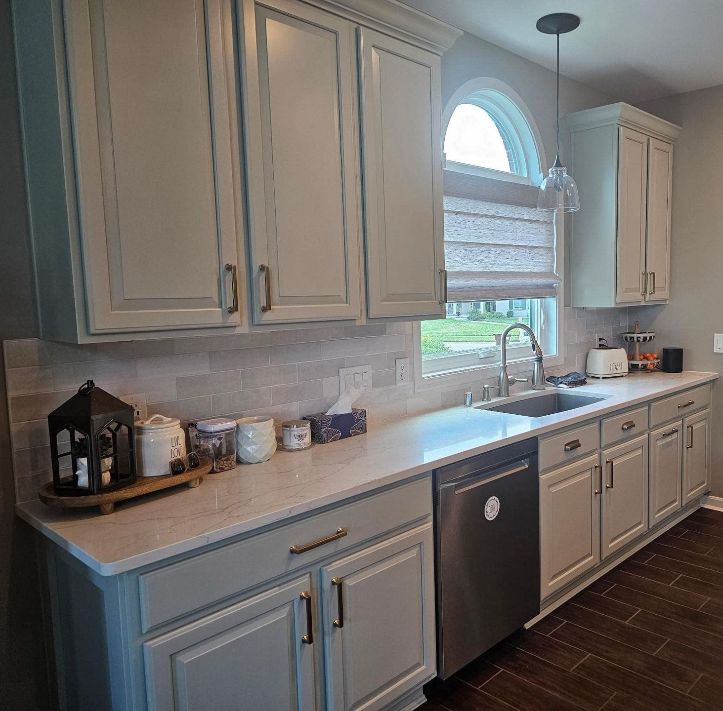 Bright kitchen with white cabinets, gray countertops, arched window, and dark dishwasher.