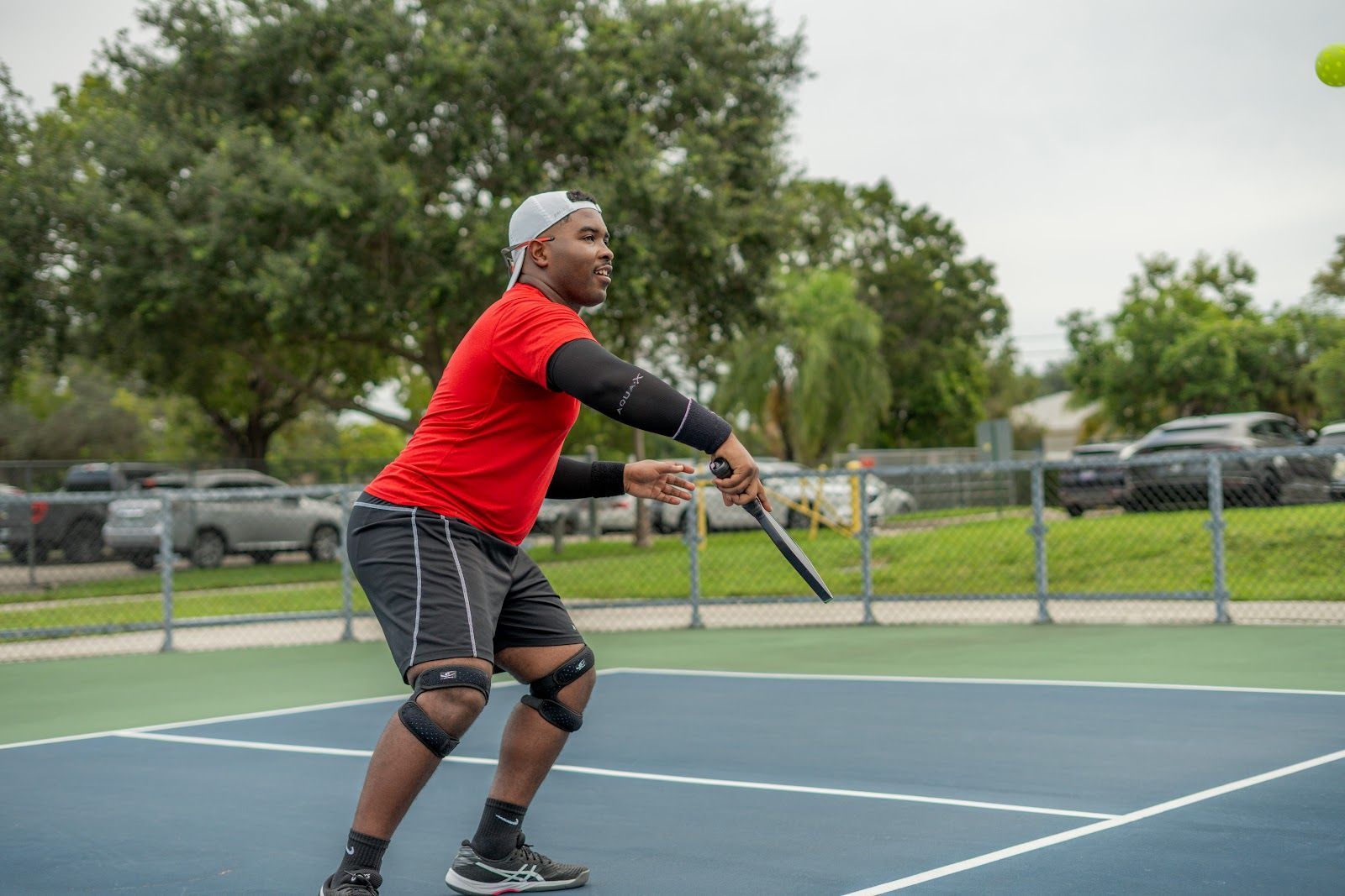 Person in red shirt and black shorts plays tennis, hitting a yellow ball on outdoor court.