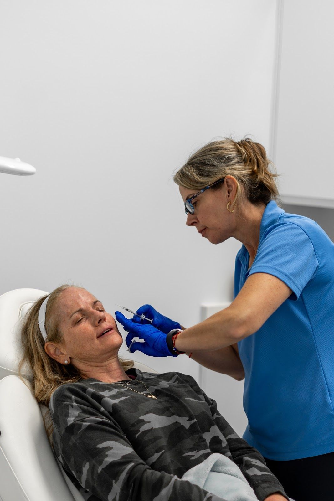 Woman receiving facial injections from a person in blue gloves and shirt. White medical room setting.