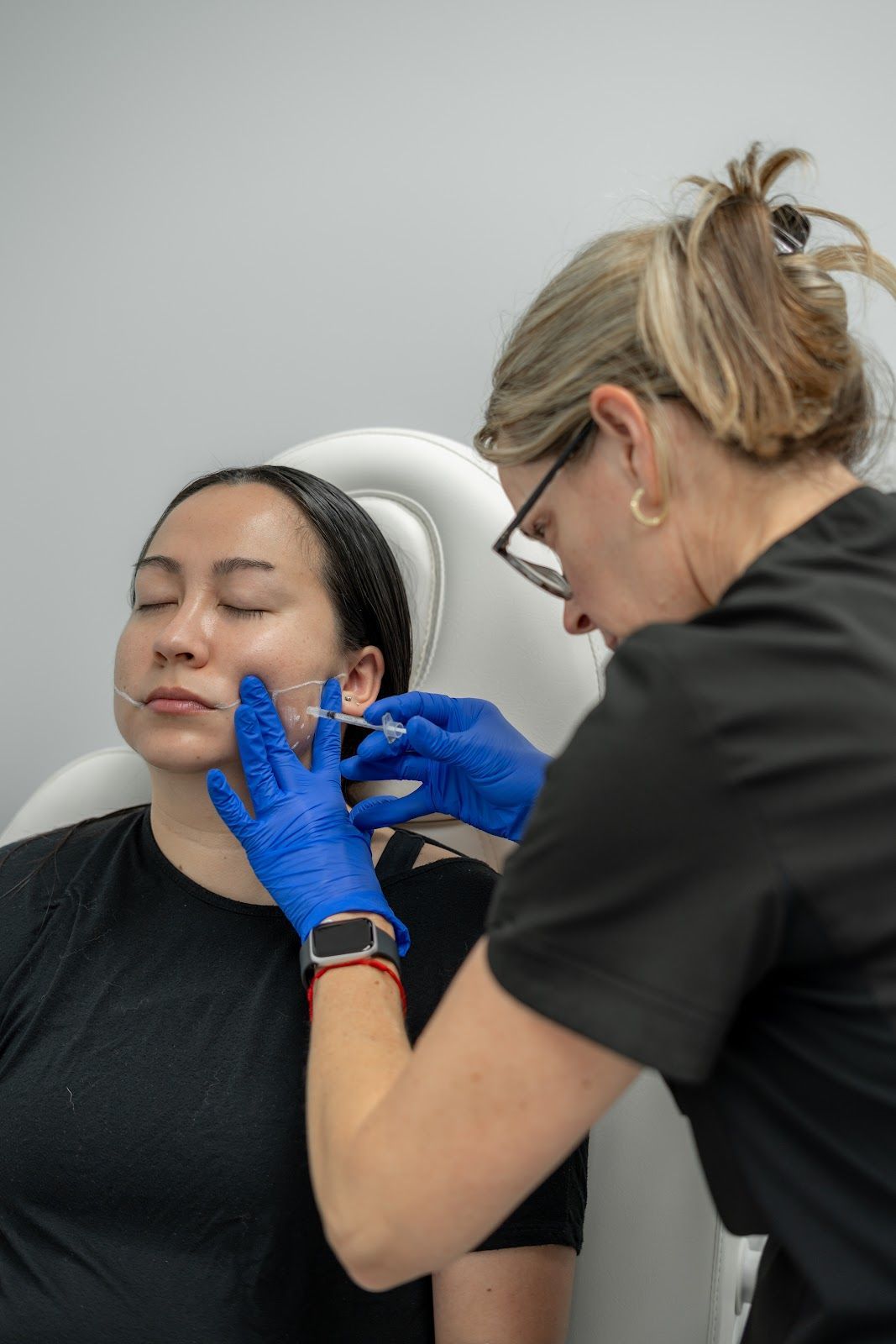 A woman receives a facial injection from a person wearing blue gloves in a clinic setting.