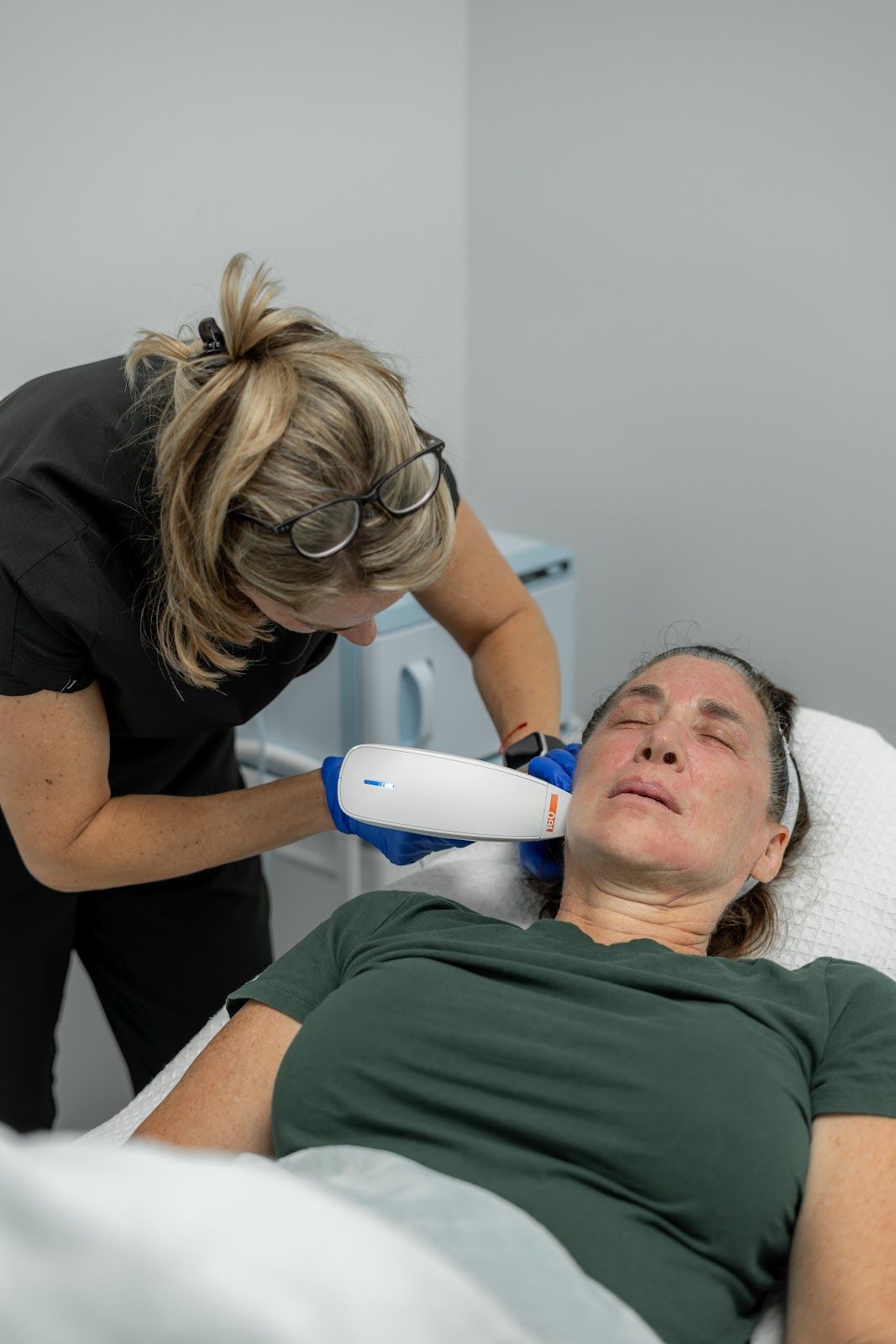 A woman receives facial treatment. A person in gloves holds a device to the woman's cheek in a medical office.