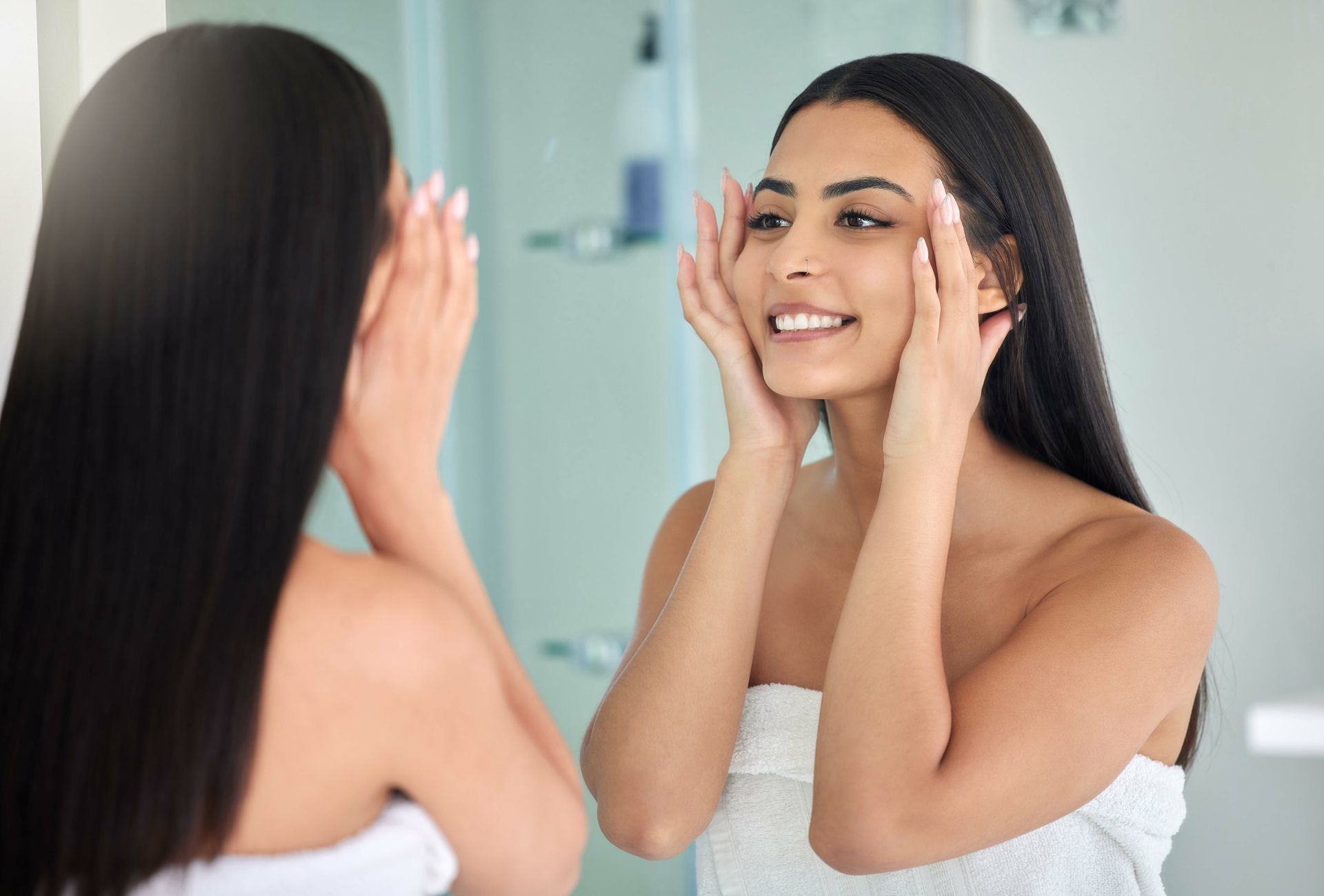 Woman in a white towel smiling at her reflection while applying skincare in a bathroom mirror
