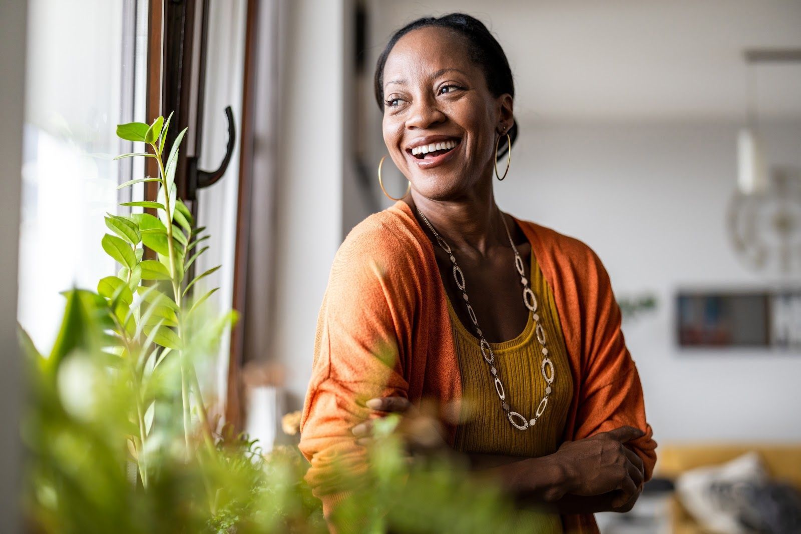 Woman smiling, looking out window, wearing orange cardigan and gold necklace, near greenery.