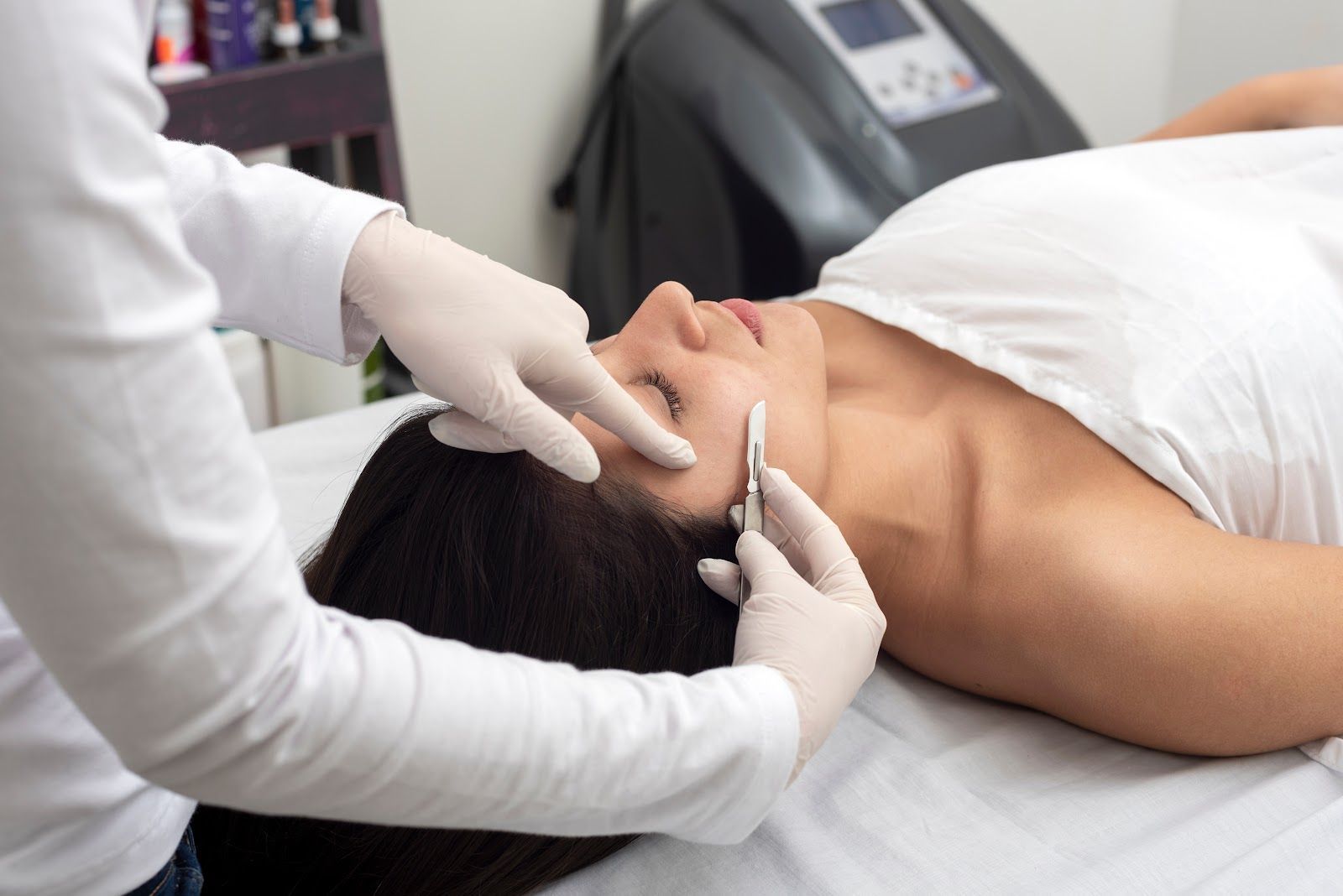 Person receiving cosmetic procedure on face, lying on a white bed. A gloved hand holds a tool near their cheek.