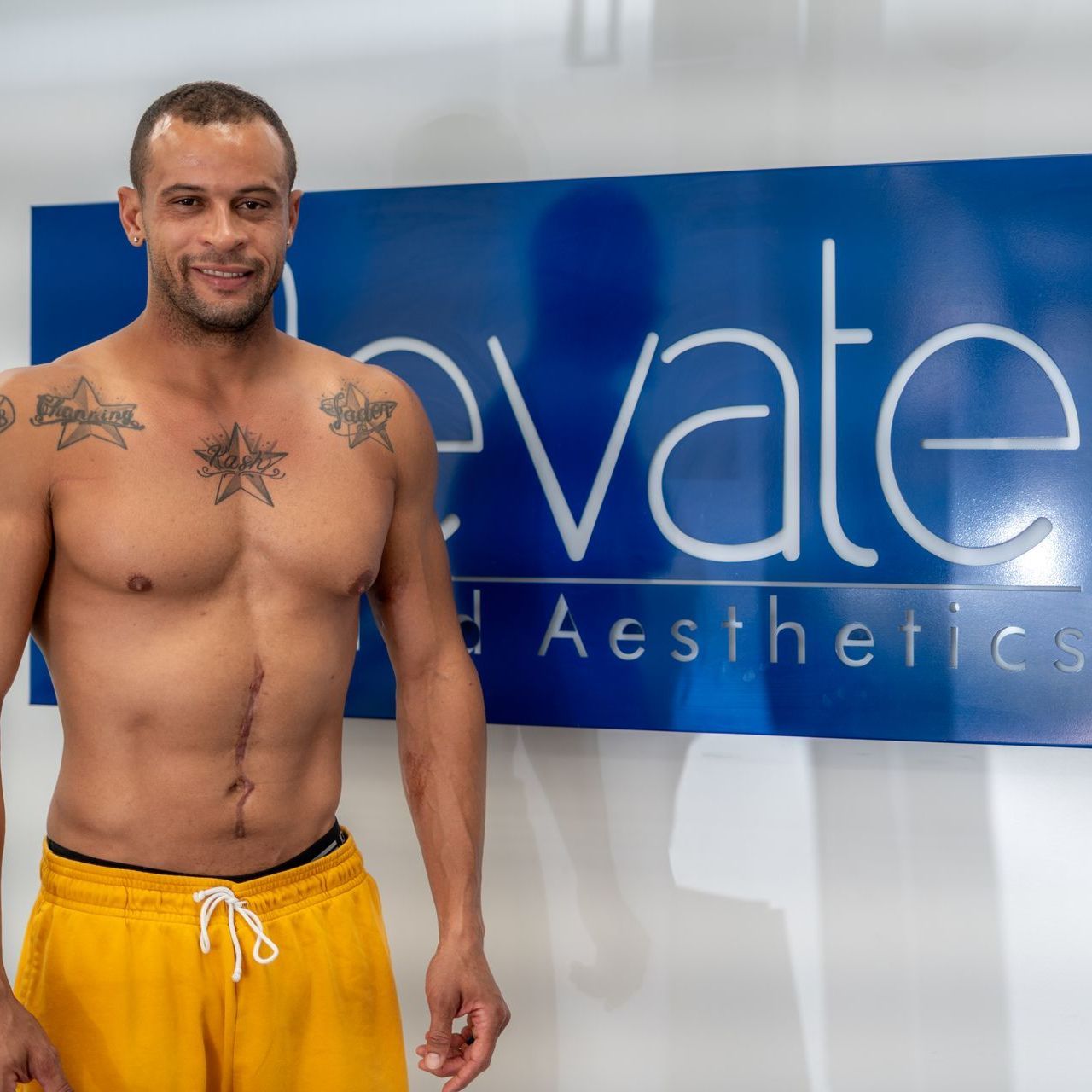 A shirtless man stands in front of a sign that says elevate aesthetics