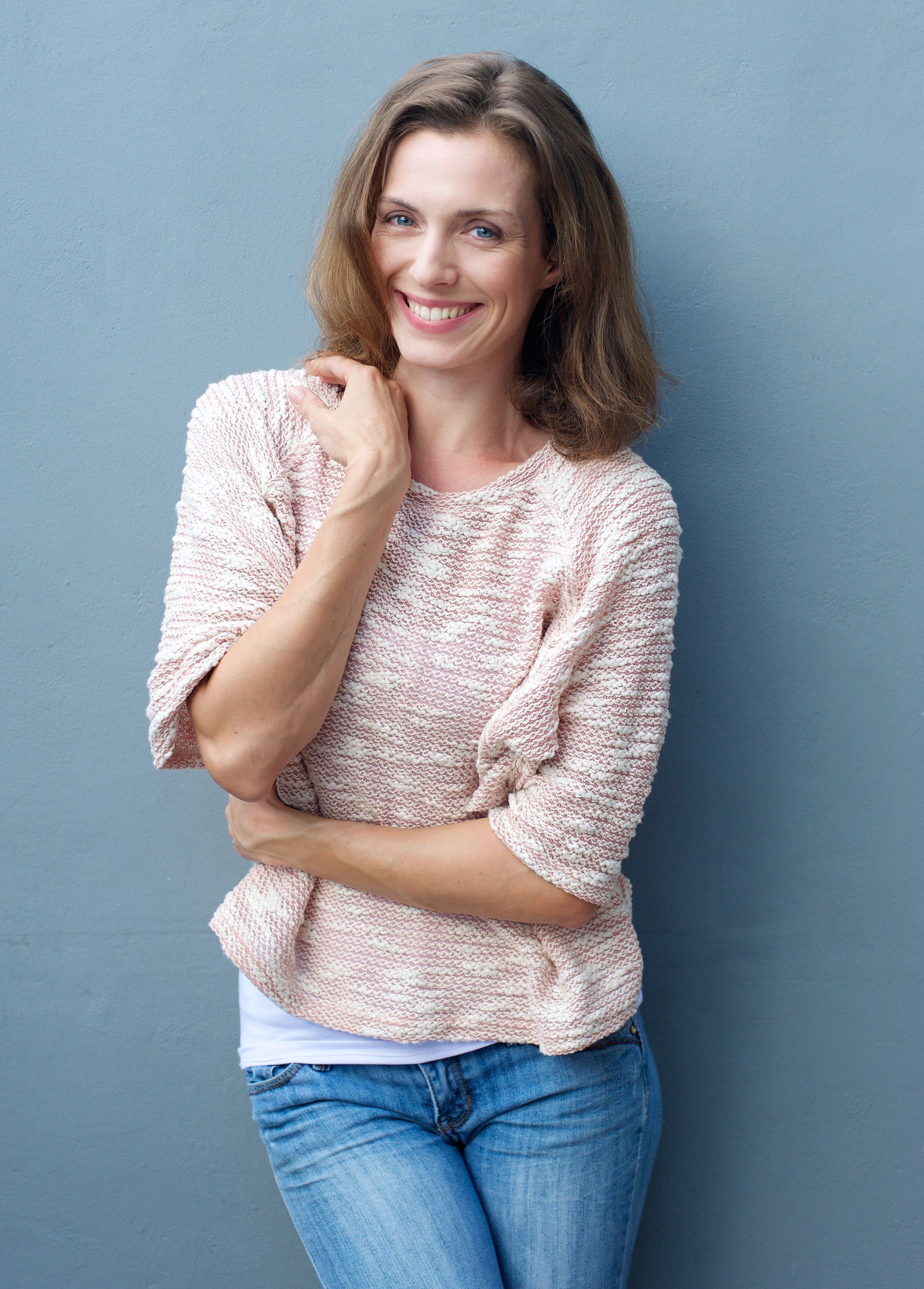 A woman in a pink sweater and blue jeans is standing in front of a gray wall.