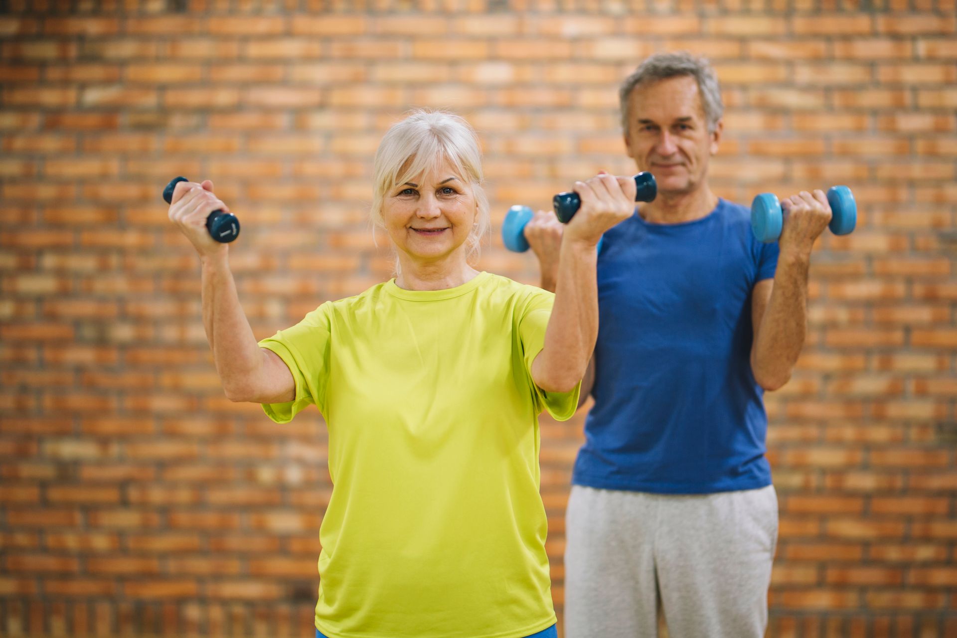 A man and a woman are holding dumbbells in front of a brick wall.