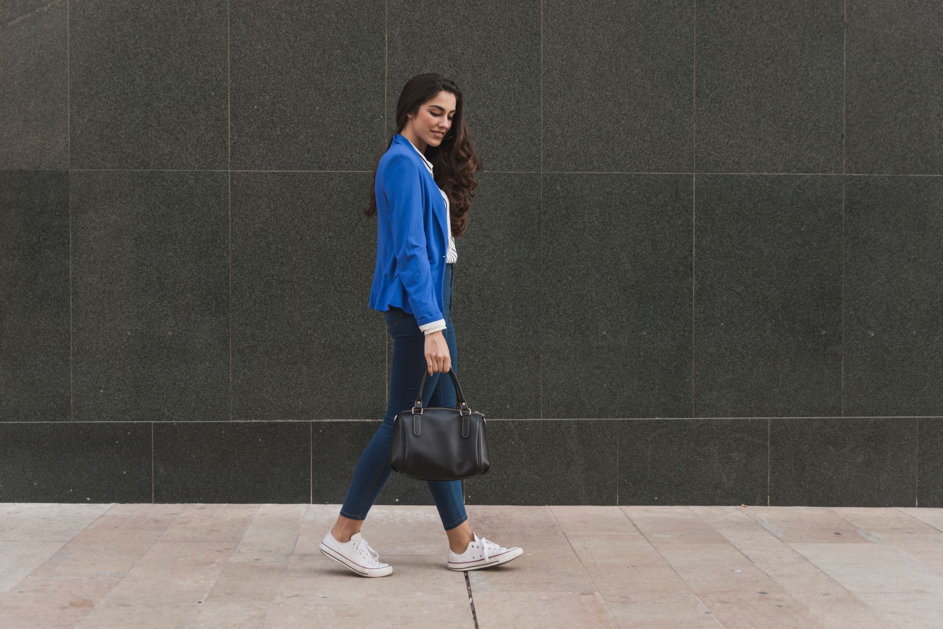 A woman in a blue jacket is walking down the street with a black purse.