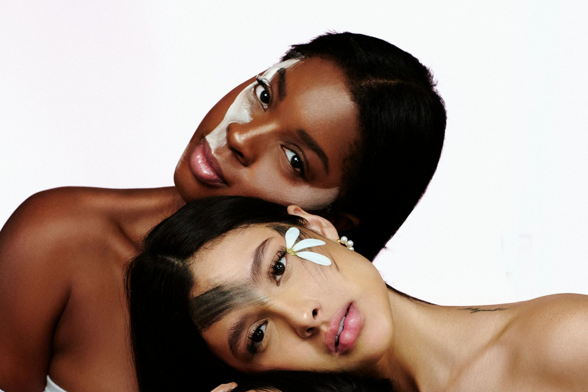 Two people with painted faces, close together against a white background. One has a floral accent, the other a mask.
