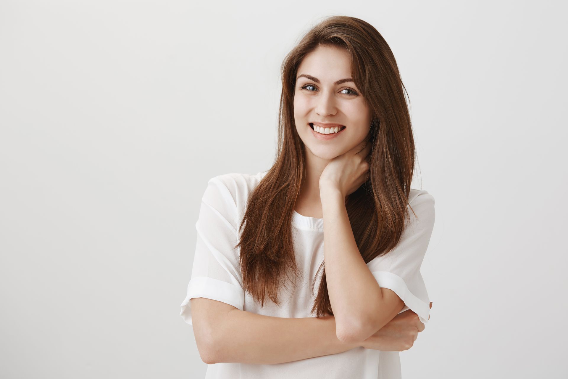 A woman in a white shirt is smiling with her arms crossed and her hand on her chin.