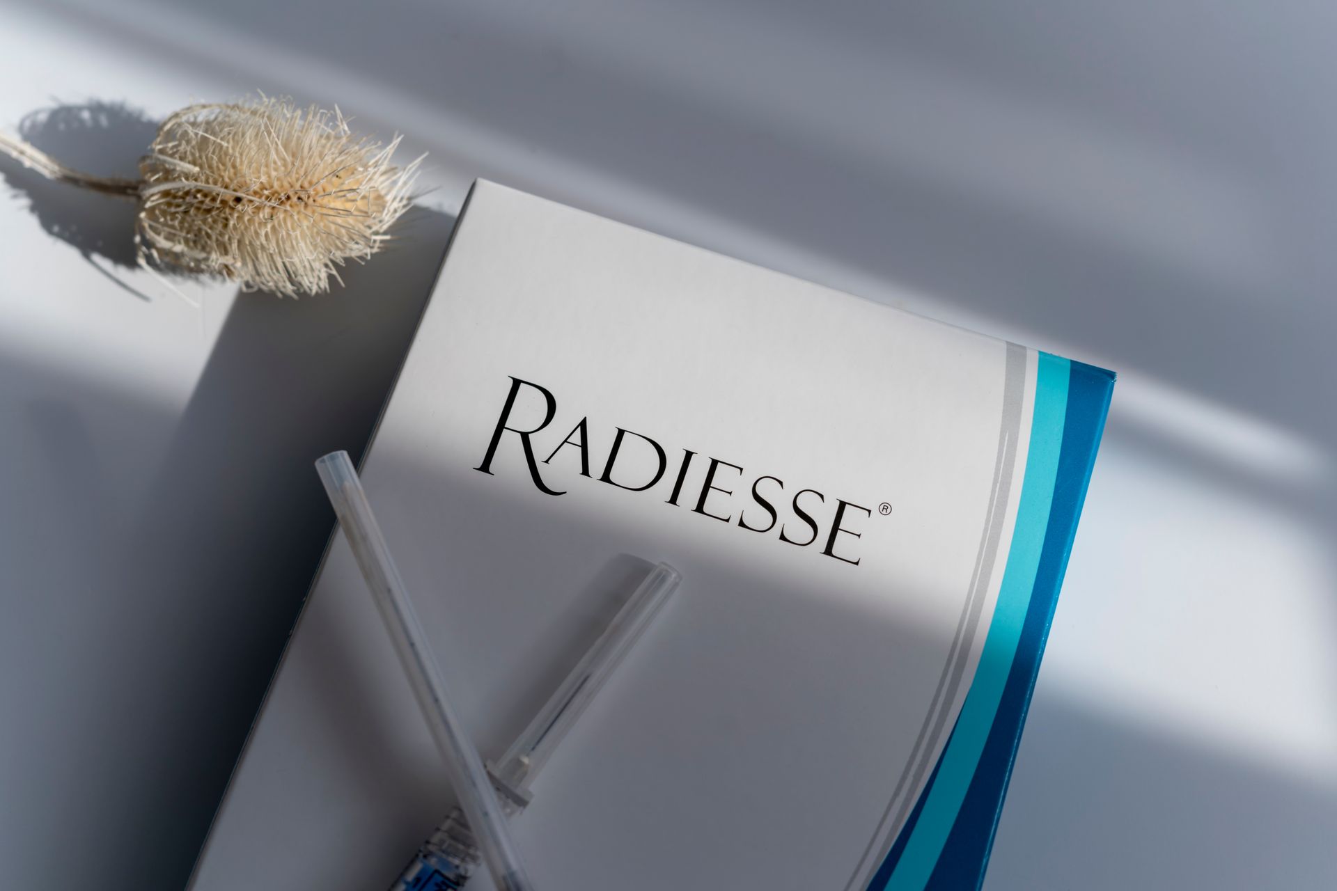 Box of Radiesse dermal filler, with syringes, and a dried flower on a white surface with shadows.