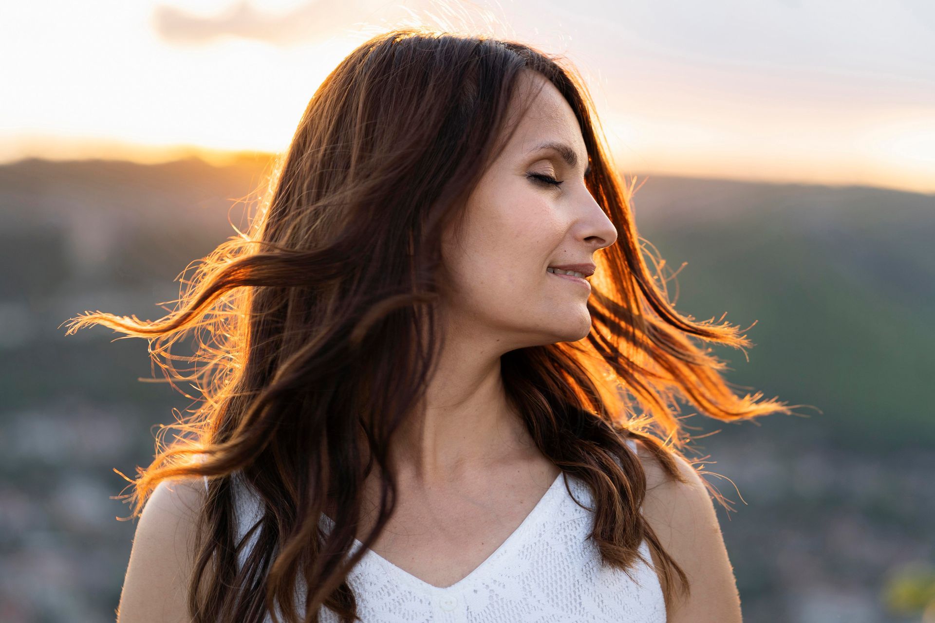 A woman in a white dress is standing on top of a hill with her eyes closed and her hair blowing in the wind.