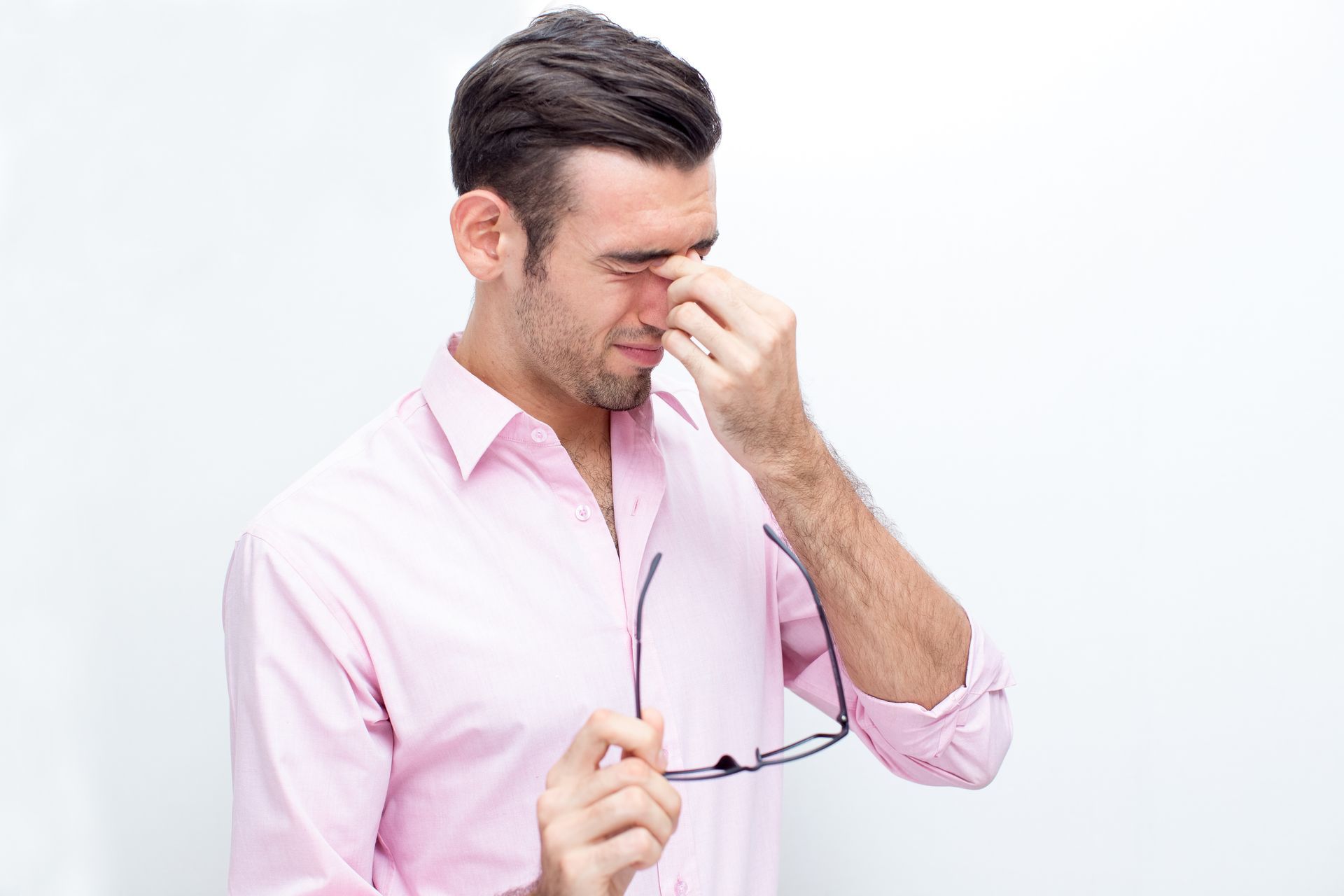 A man in a pink shirt is holding his glasses and rubbing his nose.