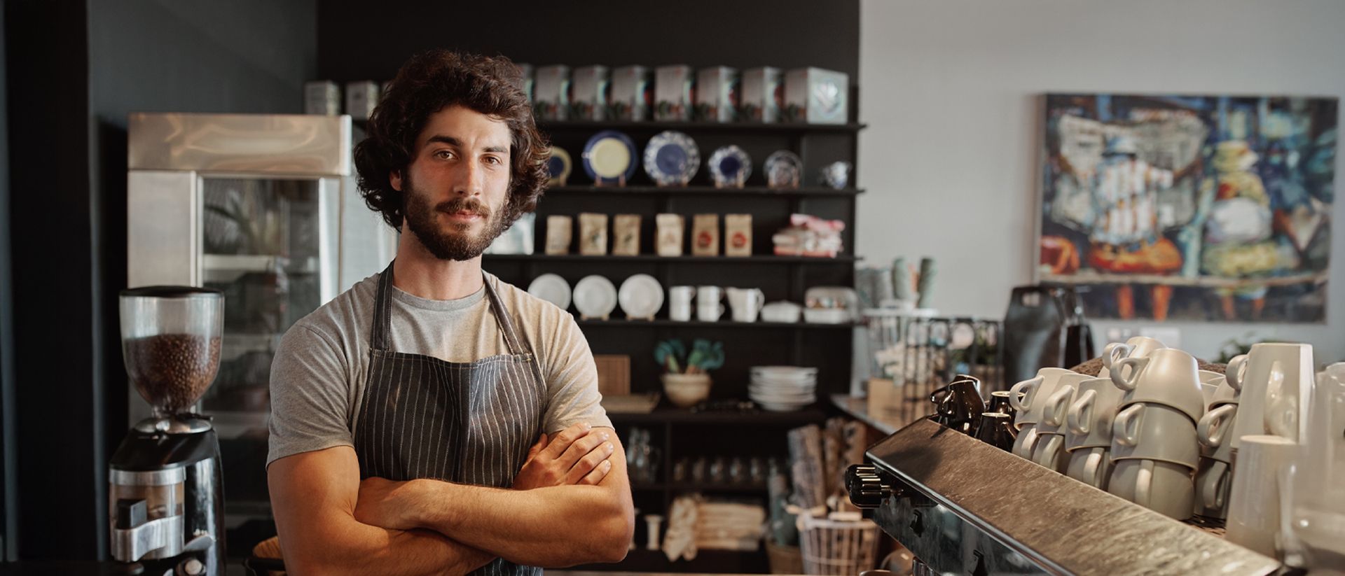 Man with curly hair, wearing an apron, standing in a coffee shop with arms crossed.
