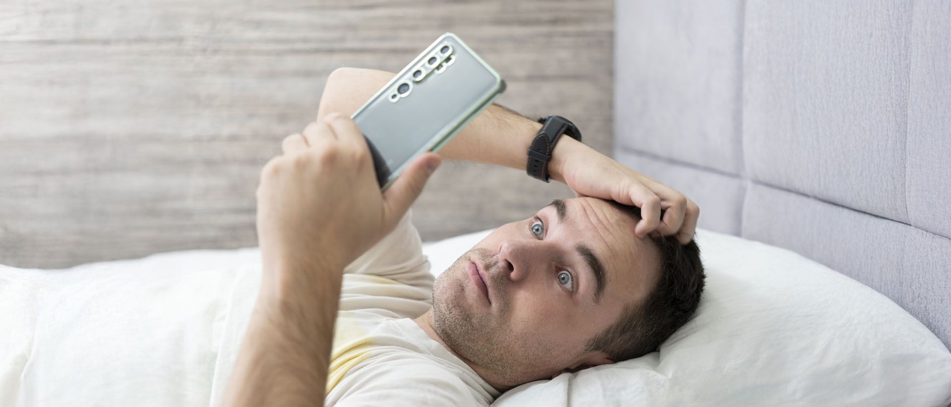 Man lying in bed, looking at his phone with a frustrated expression.